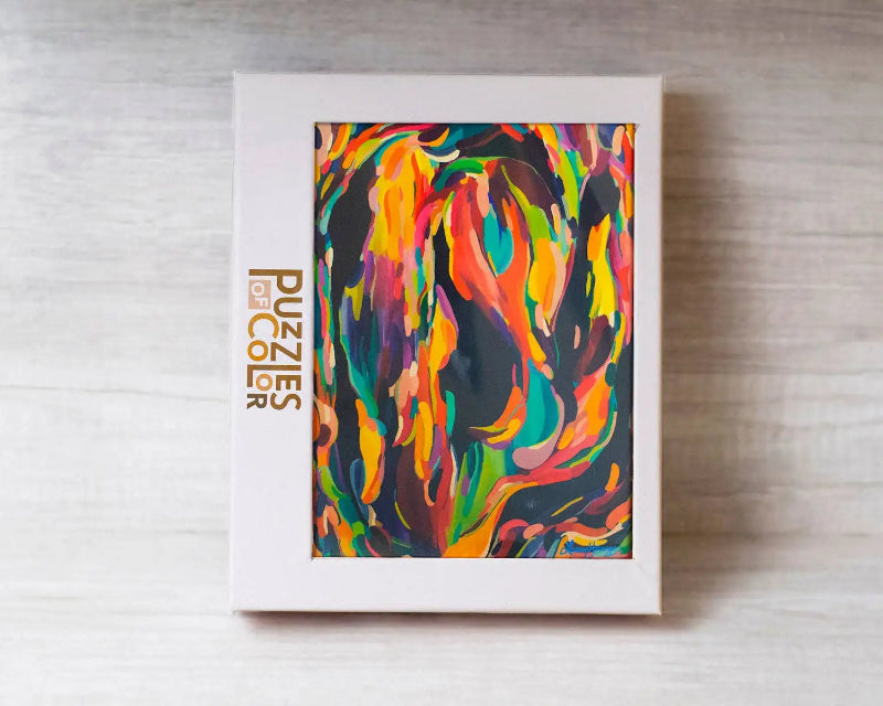 A white rectangular puzzle box standing upright on a light wood surface, featuring a colorful abstract painted design on the front and the vertical brand text 'Puzzles by Alex' in gold on the left side.