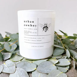 White candle with 'urban cowboy' branding and a bee illustration on a bed of green leaves.