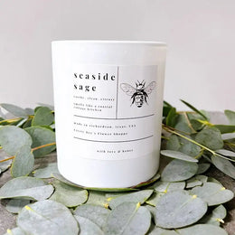 Candle labeled 'seaside sage' with a bee design on a bed of eucalyptus leaves.