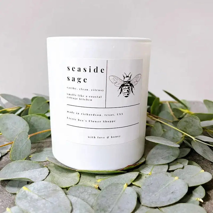 Candle labeled 'seaside sage' with a bee design on a bed of eucalyptus leaves.