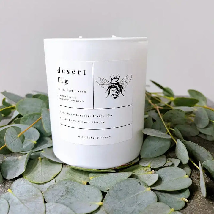 White candle with a label featuring a bee design, surrounded by eucalyptus leaves on a light gray background.