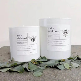 Two candles labeled 'gal's night out' with eucalyptus leaves on a concrete surface.