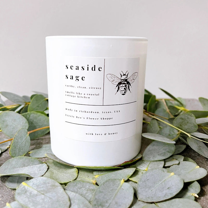 Candle labeled 'seaside sage' surrounded by eucalyptus leaves on a neutral background
