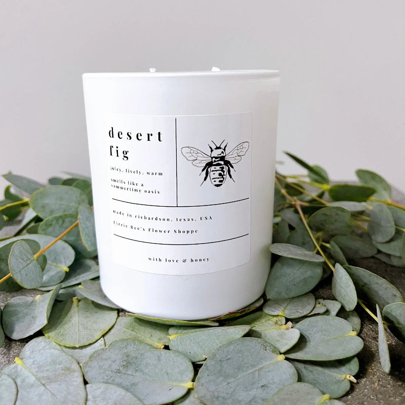 White candle with a label featuring a bee design, surrounded by eucalyptus leaves on a neutral background.