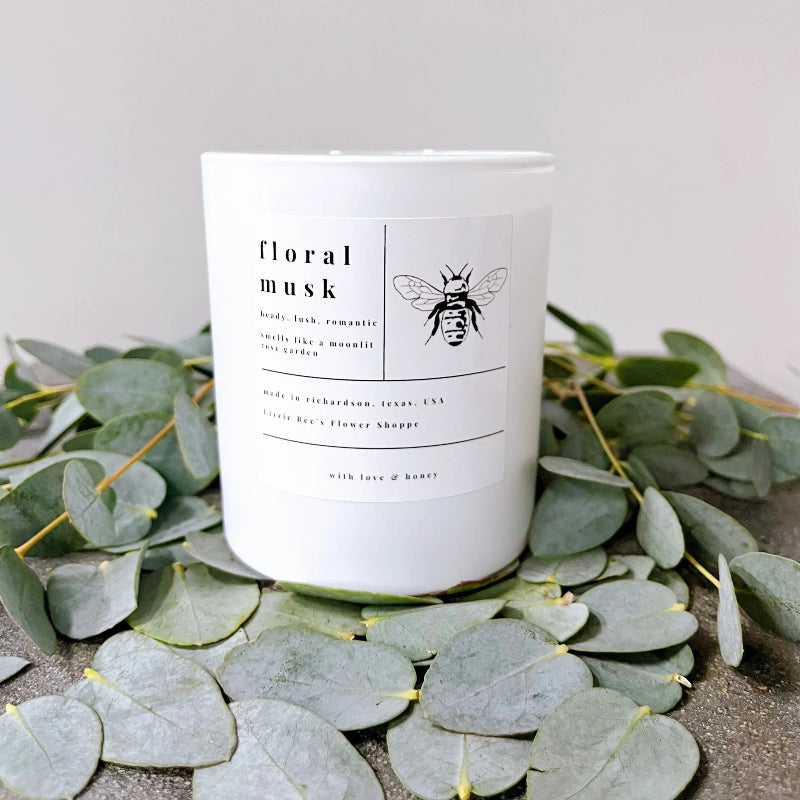Floral musk candle with a bee illustration on a bed of eucalyptus leaves.