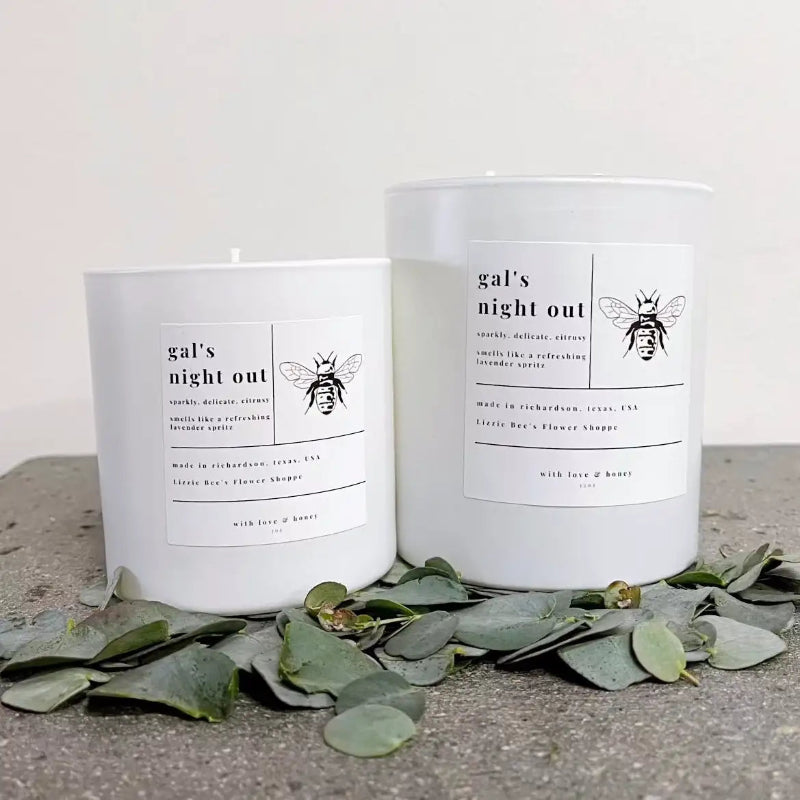 Two candles labeled 'gal's night out' with bee graphics on a concrete surface with eucalyptus leaves.