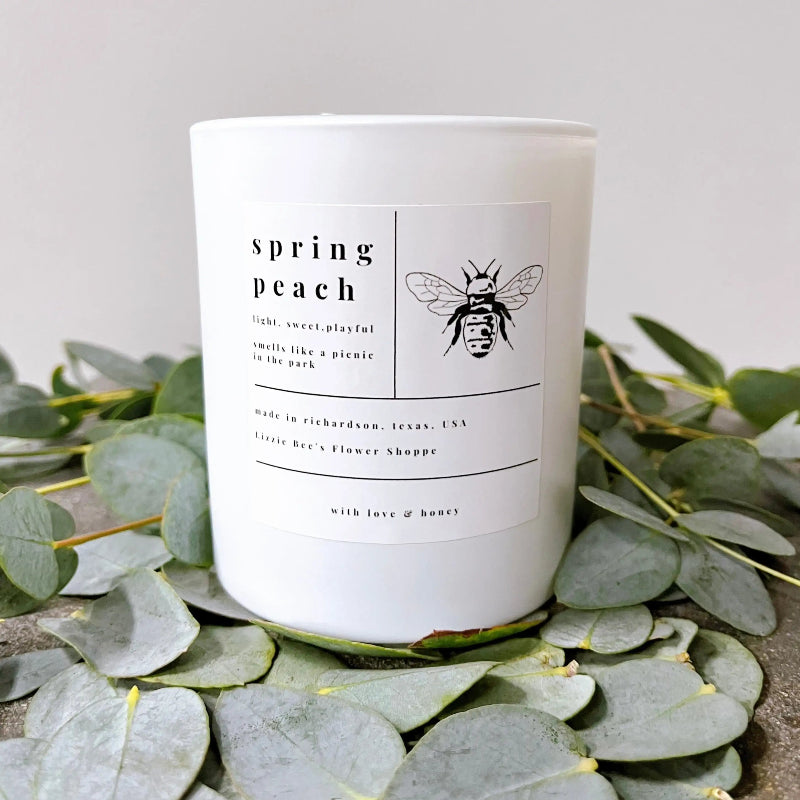 White candle labeled 'spring peach' with a bee design, placed on eucalyptus leaves.