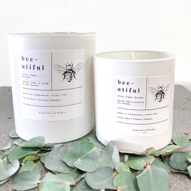 Two white candles with bee-themed labels on a surface with green leaves.