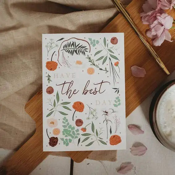 Card with floral design and text 'Have the best day' on a wooden surface with flowers and a cup.