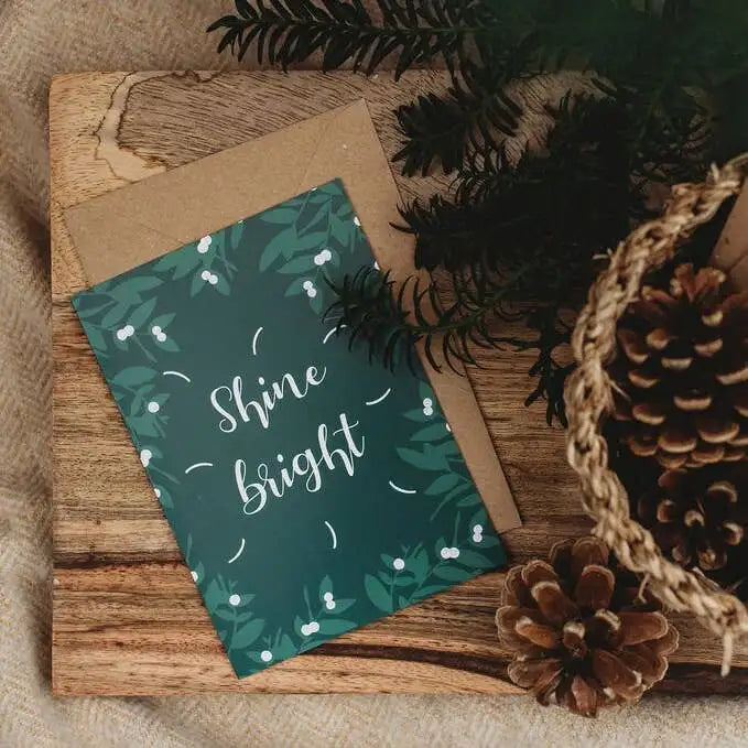Greeting card with 'Shine bright' text on a wooden surface with pinecones and branches.