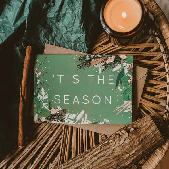 Card with 'Tis the Season' text on a decorative setting with a candle and greenery.