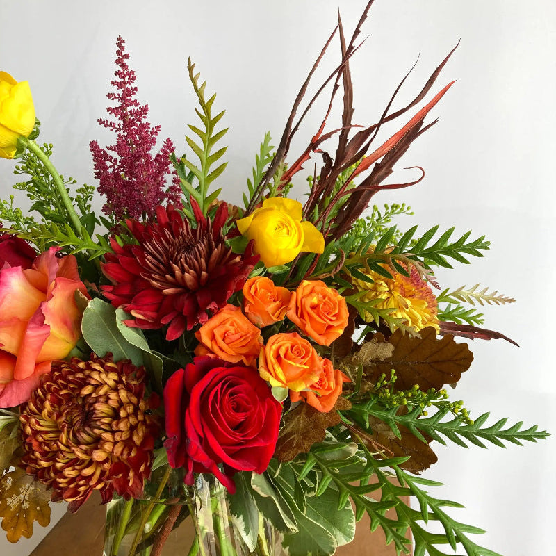 Bouquet of flowers with red, orange, and yellow roses in a clear vase on a wooden surface.