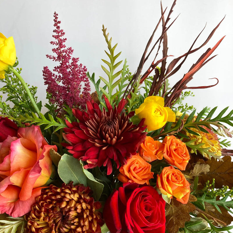 Bouquet of colorful flowers including red roses, yellow and orange roses, and greenery on a light background.