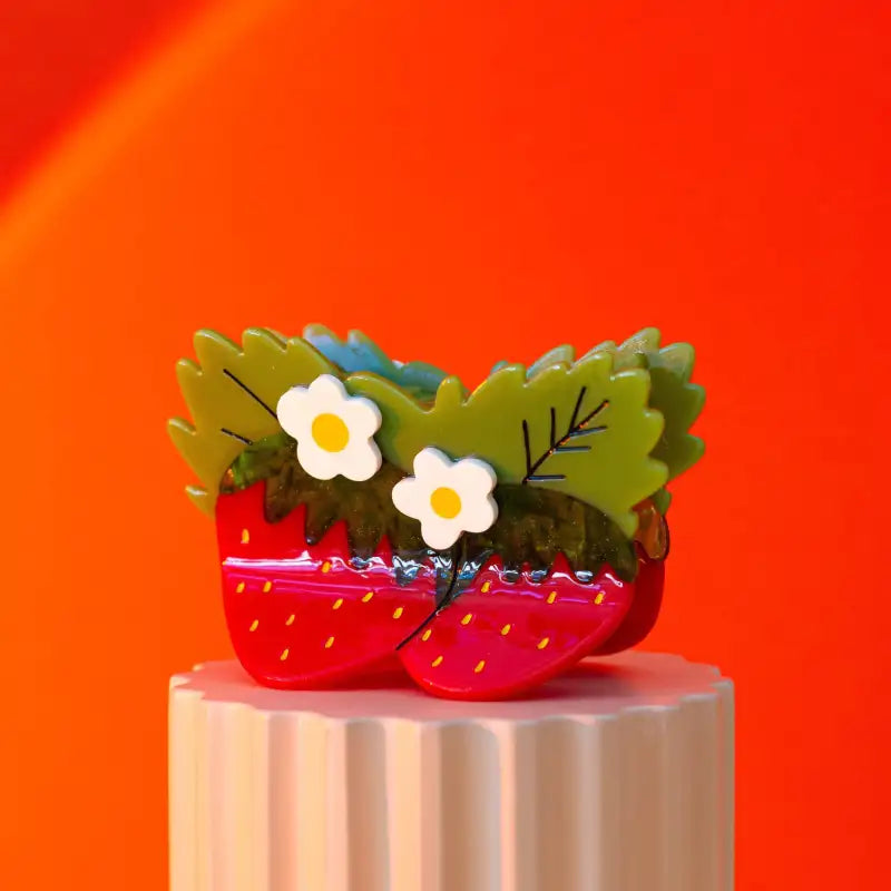Strawberry-shaped hair claw on a pink platform against an orange background