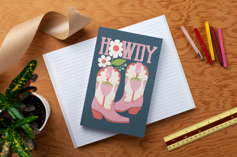 Notebook with a 'Howdy' card featuring cowboy boots and flowers on a wooden surface.