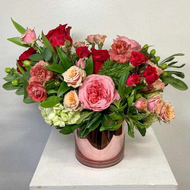 Bouquet of red, pink, and green flowers in a pink vase on a white surface.