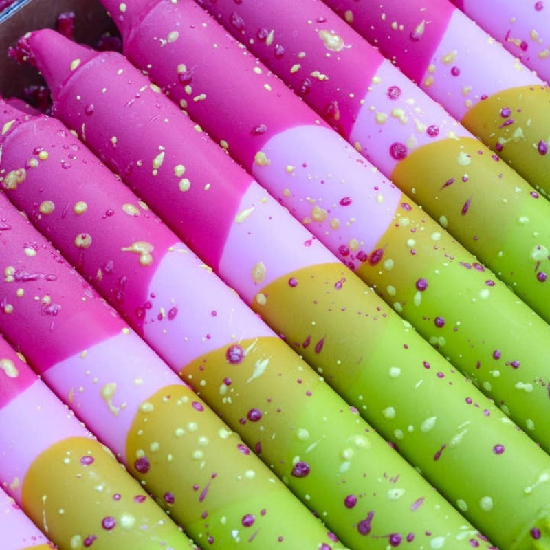A close-up of several taper candles showing color-blocked sections of bright pink, light lavender, and lime green, with scattered confetti and small purple and white speckles.