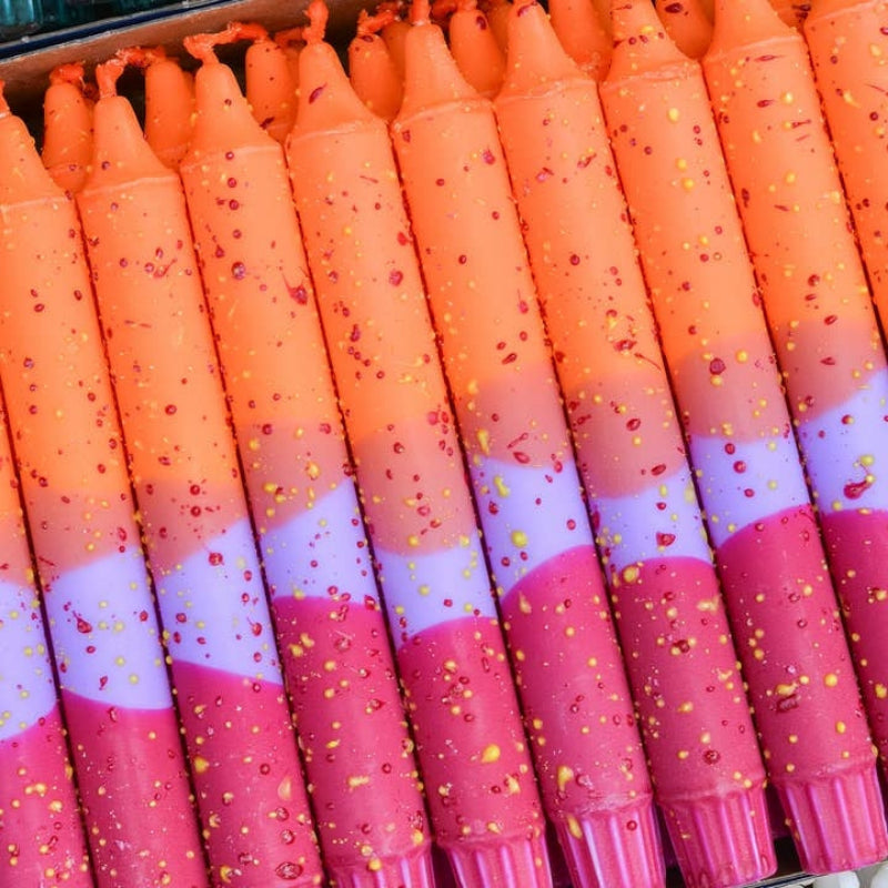 A close-up of multiple orange taper candles arranged side by side, each with a pink lower section and scattered small red and yellow confetti speckles.