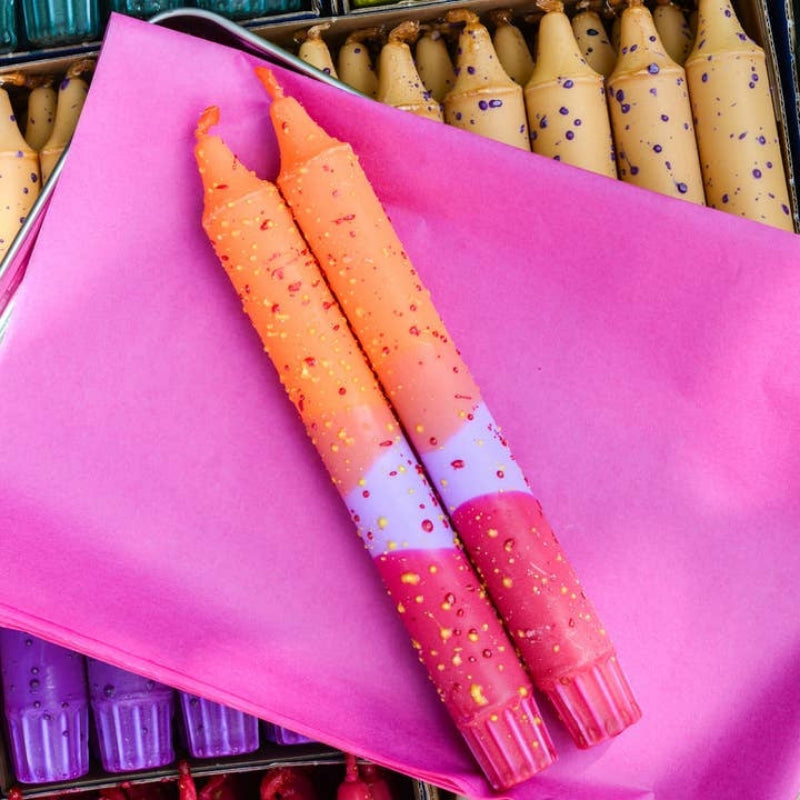 Two decorative taper candles with a pink, purple, and orange gradient design and confetti speckles, resting on a bright pink sheet of paper inside a metal tray with other candles visible in the background.