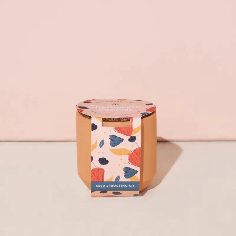 A small, cylindrical, light brown terracotta Modern Sprout Tiny Terracotta Grow Kit, Champagne Poppies variety, featuring a colorful abstract floral label and small text reading 