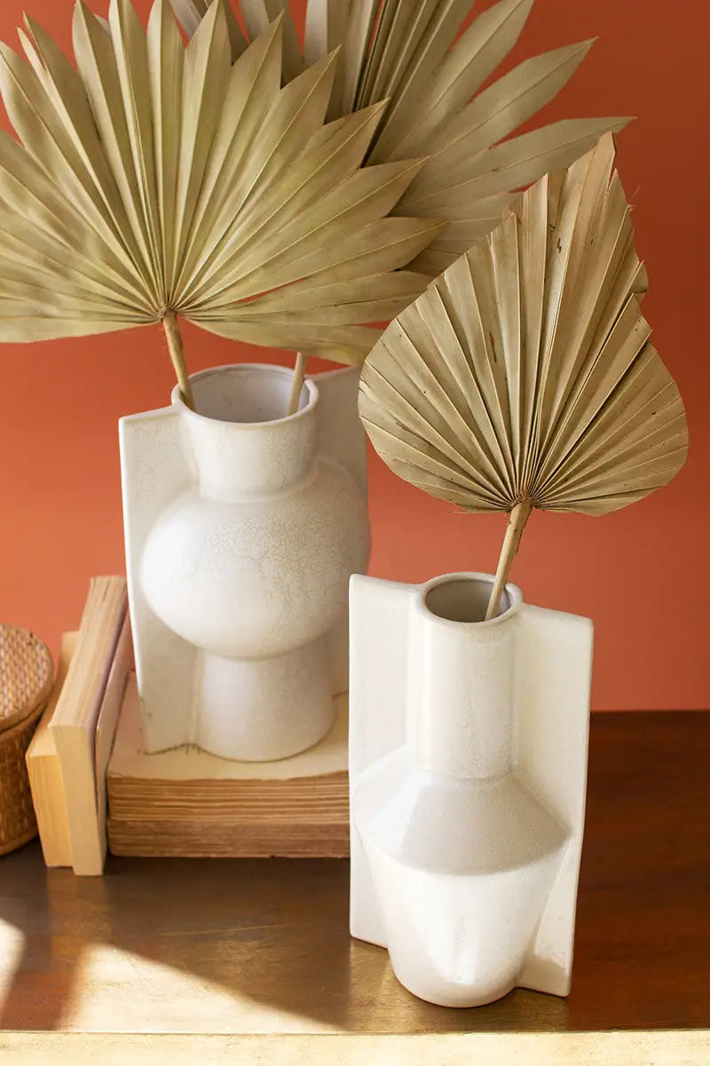 Two white vases with dried palm leaves on a wooden surface against an orange wall.