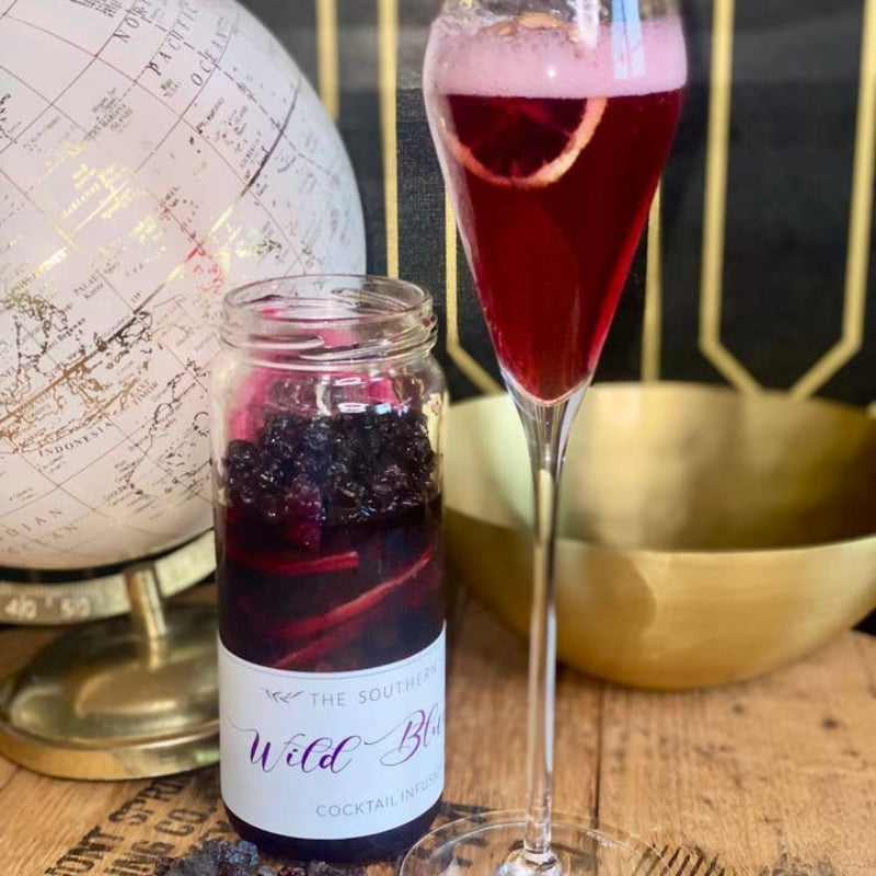 A glass jar candle labeled 'The Southern Wild Berry Cocktail Infused' next to a filled champagne flute with a berry garnish, styled on a wooden surface with a globe and gold bowl in the background.