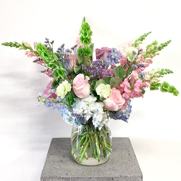 A floral arrangement featuring a variety of flowers including roses and other greenery, in pastel colors, placed in a tall clear vase.