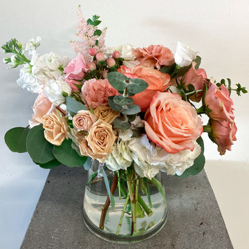 A floral arrangement featuring a collection of classic blooms in white, blush, and peach tones, in a clear glass vase.